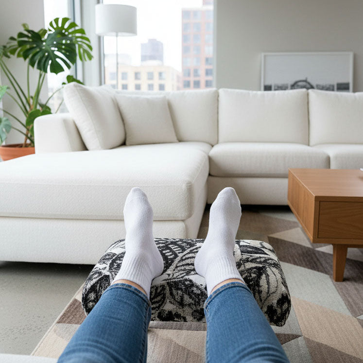 Handcrafted small footstool. It has a black and white leaf pattern fabric top. It is in a modern living room. Someone has their feet up on it. Made by Little Forest Studio.
